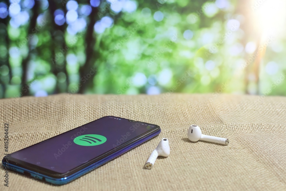 Kedah, Malaysia- 1st February 2021: Selective focus image of smartphone showing Spotify icon logo and wireless earphones. Bokeh effect background